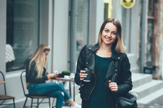 Take away coffee. Beautiful young urban woman wearing in stylish clothes holding coffee cup and smiling while walking along the street. Student's coffee break after study. Fashion lifestyle.