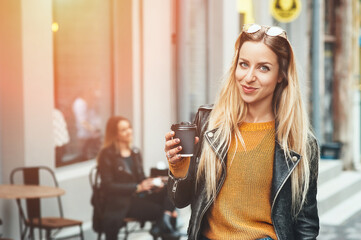 Take away coffee. Beautiful young urban woman wearing in stylish clothes holding coffee cup and smiling while walking along the street. Student's coffee break after study. Fashion lifestyle.