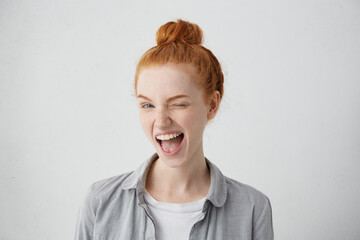 Funny red-haired female teenager with bun wearing casual shirt having joyful expression closing one...