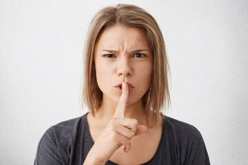 Be silent! Beautiful young Caucasian woman with stylish hairdo dressed casually holding finger at her lips asking to be quiet having angry expression. Secrecy and concealing important information