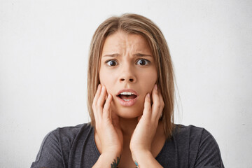 Young woman looking with terror into camera holding her hands on cheeks isolated over white background. Pretty girl having despair looking with wide opened eyes and mouth shouting loudly with panic