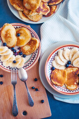 Homemade pancakes with banana and black currant on painted ceramic plates, with wooden board and tablecloth, with vintage fork and spoon. Flatlay food photo.