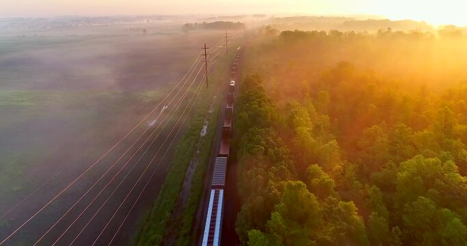 Freight train rolls through foggy rural landscape at sunrise, aerial view, breathtaking scenic beauty.
