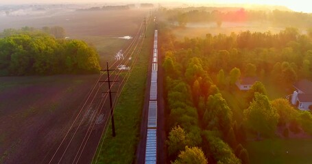 Freight train rolls through foggy rural landscape at sunrise, aerial view, breathtaking scenic beauty.
