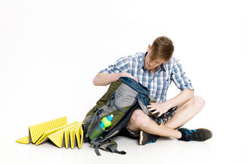 Young tourist is sitting and looking in backpack something on white background. traveler is...