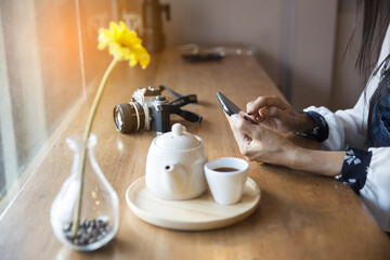 Shopping and relaxing concept,woman using smartphone to shopping .Beautiful girl sending message to friends or boyfriend at coffee shop.Professional camera,tea set and vase of flower on the desk.