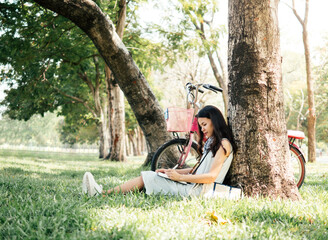 asian woman smiling and  sitting under a tree reading & writing a diary book in the park, with bicycle