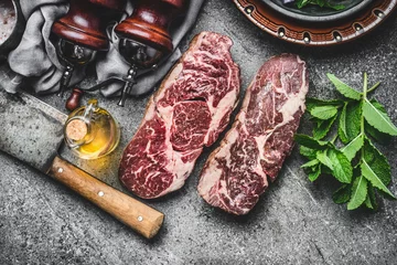 Photo sur Plexiglas Viande Two Dry aged raw beef steaks with meat cleaver and condiment on dark rustic concrete background , top view
