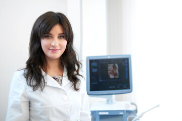 Portrait of professional female nurse in white uniform at modern and new clinic, standing at cabinet, posing, smiling at camera. Attractive brunette doctor against ultrasound equipment and computer.