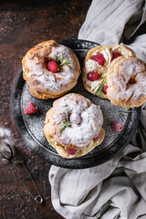Homemade choux pastry cake Paris Brest with raspberries, almond, sugar powder and rosemary, served on black wooden serving board over dark texture background with textile. French dessert. Top view