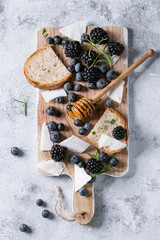 Berries blackberry and blueberry, honey on dipper, rosemary, sliced goat cheese with bread served on wooden board over gray texture background. Summer sandwich. Top view with space