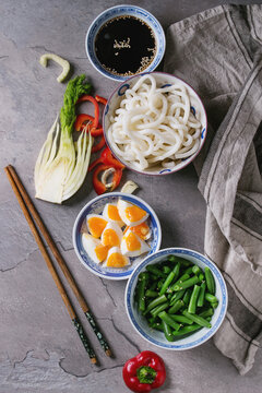 Ingredients for cooking stir fry udon noodles, green beans, sliced paprika, boiled eggs, soy sauce with sesame seeds in traditional bowls with wooden chopsticks over gray texture background. Flat lay