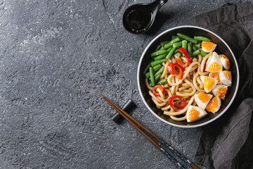 Cooking stir fry udon noodles, green beans, sliced paprika, boiled eggs, soy sauce with sesame seeds in black plate with wood chopsticks over black texture background. Flat lay. Asian style dinner