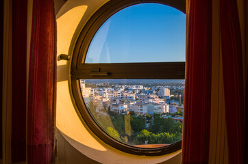 Okrągłe okno, widać przez nie panoramę miasta we wschodzącym słońcu. 