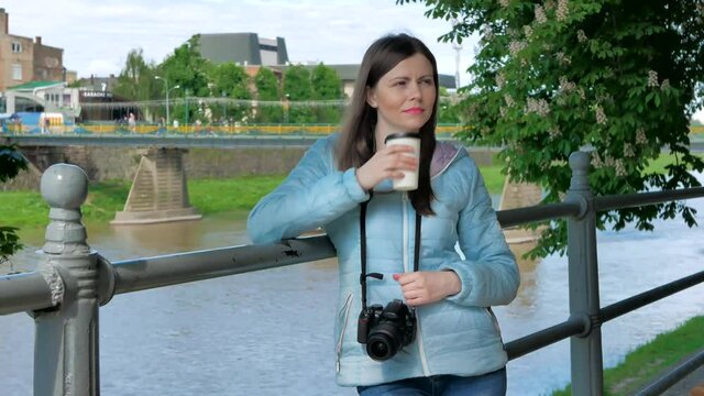 Pretty young girl photographer with a professional camera drinking coffee near the railing on waterfront, river and bridge in the background