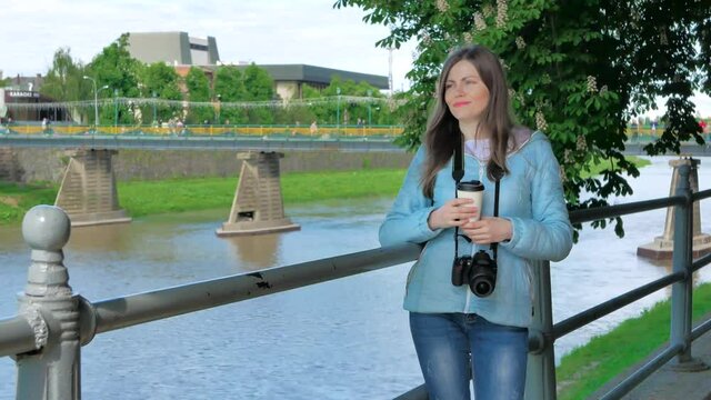 Pretty young girl photographer with a professional camera drinking coffee near the railing on waterfront, river and bridge in the background