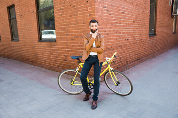 Young bearded man holding hand on chin and looking at camera while sitting near his bicycle outdoors near brick wall