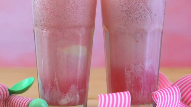 Making ice cream soda floats with pink raspberry soda and ice cream in tall cafe glasses against a pink theme summer party backgroud, slow pan up macro closeup.