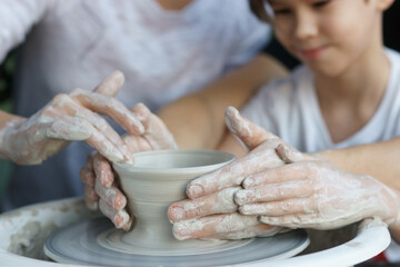 Parents teach their child to work on potter wheel. Father showing how to sculp clay pot. Closeup of dirty hands. Shallow DOF.