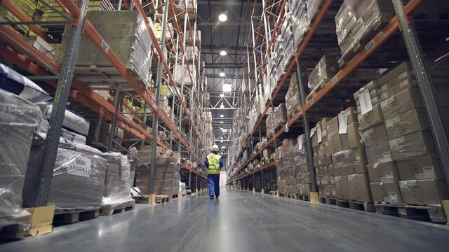 Back view. Female warehouse worker walks through rows of storage racks with merchandise