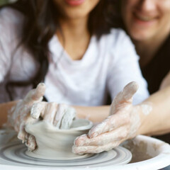 Happy smiling joyful romantic couple working together on potter wheel and sculpting clay pot. Focus on dirty hands