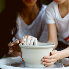 Mother teaches son to work on pottery wheel. Close up of dirty hands sculpting clay.