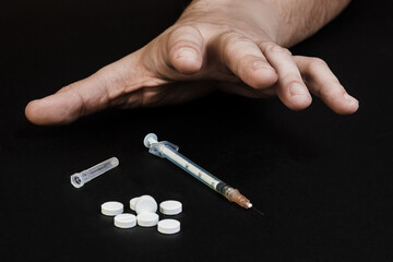 Man's hand, the addict reaches for a syringe and pills. On a black background.