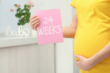 Young pregnant woman holding card with text 24 WEEKS near belly at home