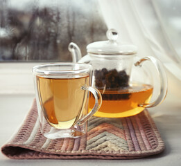Glass cup of hot tasty drink with teapot and napkin on windowsill
