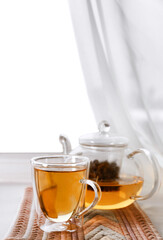 Glass cup of hot tasty drink with teapot and napkin on windowsill