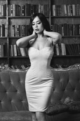 Sexy tanned brunette girl with long curly hair in beige strapless cocktail dress standing in luxury library interior. Fashion portrait of young woman with hands on her neck.