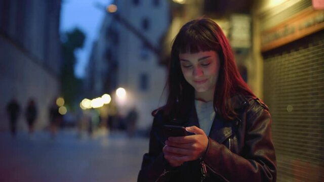 Beautiful brunette girl in leather biker jacket uses her smartphone on night street, smiling and texting