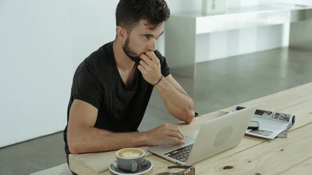 Young attractive bearded man sits in coworking place alone, works on his laptop next to cappuccino cup and suspiciously looking around, waiting his partner to come for meeting