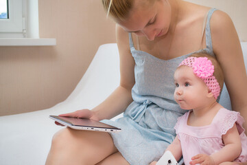 Young beautiful blonde mom and her adorable little daughter dressed in dresses are sitting on a white fluffy sofa and are teaching the use of gadgets. Modern technologies © silentalex88