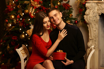 Young romantic couple sitting and hugging in christmas decorated home interior. Pretty woman in red dress with gift and man in black