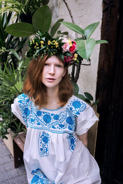 Cute young woman model with red hair and flower wreath wears handmade long white dress with ethnic flower embroidery and walks in european town on little street with plants and wooden brown old doors.