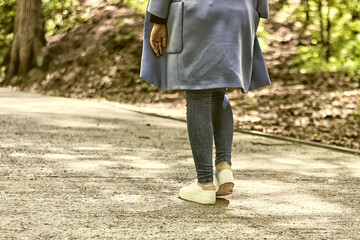 Girl walking away through alley with park at the sunny day. white sneakers and a blue coat, a spring outing.