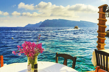 Santorini island, Oia village, Greece. Vase of pink flowers on table with white tablecloth against turquoise water of Aegean sea and mountains in background. Romantic background, restaurant scene.