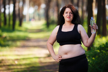 Young overweight smiling woman with a bottle of clear water outdoors