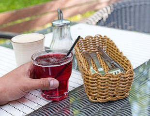 Obraz na płótnie Canvas cherry juice in the male hand with black tube, glass Cup, basket with about Cutlery, coffee Cup, sugar bowl, table
