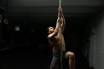 Male athlete or sportsman climbing rope at dark gym. Topless handsome muscular man in gray shorts.
