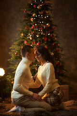 Young romantic couple sitting on floor in christmas decorated interior against christmas tree
