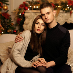 Fototapeta premium Cute beautiful caucasian young couple sutting at sofa in christmas decorated interior and hugging. Romantic couple staying at home and enjoying tome together