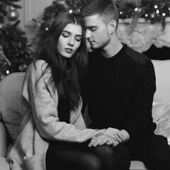 Cute beautiful caucasian young couple sutting at sofa in christmas decorated interior and hugging. Romantic couple staying at home and enjoying tome together