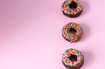 Three baked chocolate doughnuts with chocolate glaze with confetti over pink background. Overhead view.