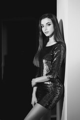 Fototapeta premium Young sexy girl wearing blue green shiny dress with sequins standing in inreior near wall. Fashion model with long brunette hair and makeup