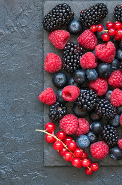 Mix of berries raspberries red currants and blueberries on black slate board. Gray stone background.  Top view.