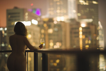 Beautiful naked female on a balcony with a glass of white wine on a background of a night city in...