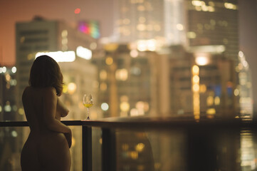 Beautiful naked female on a balcony with a glass of white wine on a background of a night city in Kuala Lumpur. Malaysia