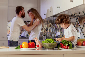 Small boy with the phone in his hands sitting on the kitchen surface while his parents fighting on the background.